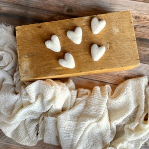 May include: Five white felt hearts arranged on a wooden box with a white fabric draped over the box.