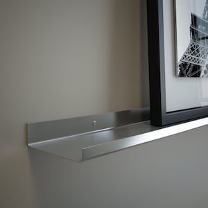 May include: A silver metal shelf with a slightly curved edge. The shelf is mounted to a wall and has a picture frame resting on it.