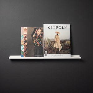 May include: Two magazines on a white shelf against a black wall. The magazine on the left has a woman wearing a floral top and black skirt. The magazine on the right is titled "KINFOLK" and features a woman wearing a straw hat and a long white dress.