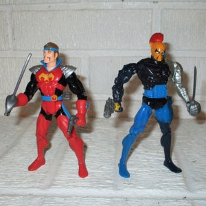 May include: Two action figures, one in red and black with a sword and gun, the other in black and blue with a sword and gun. The figures have detailed armor and are posed against a white brick background.