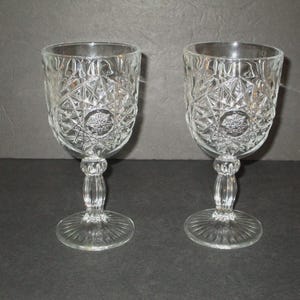 May include: Two clear crystal wine glasses with intricate diamond and starburst patterns. The glasses have a round bowl, a decorative stem, and a circular base. The glasses are displayed on a dark surface.