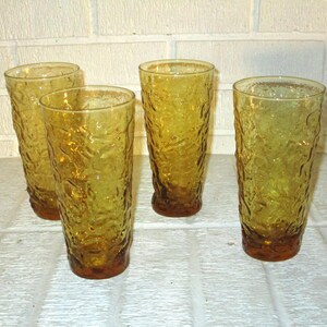 May include: Four amber-colored drinking glasses with a textured, organic pattern. The glasses are tall and cylindrical, with a slightly darker amber base. They are arranged on a white tiled surface.