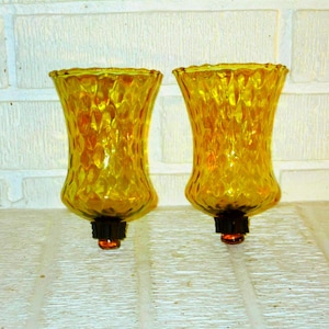 May include: Two amber-colored glass candle holders featuring a textured, diamond-patterned surface. Each vessel has a flared rim and tapers toward a dark, circular base with a small, protruding amber glass knob at the very bottom center.