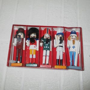 May include: A set of five wooden nutcracker figurines in a red display box. The figures include a golfer in brown, a soccer player with number 10, a football player with number 25, a baseball player in pinstripes, and a skier in white and blue.