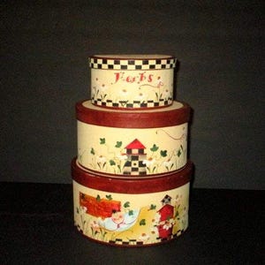 May include: A stack of three round, decorative boxes with lids. The boxes are painted with a cream-colored background and feature a red rim and a black and white checkered pattern on the lids. The top box says "Herbs".