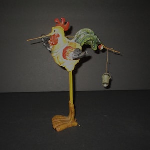 May include: A decorative rooster figurine with a yellow body, red comb, and green tail feathers. The rooster stands on a single long yellow leg and carries a thin wire across its shoulders, from which a small metal bucket hangs on one side.