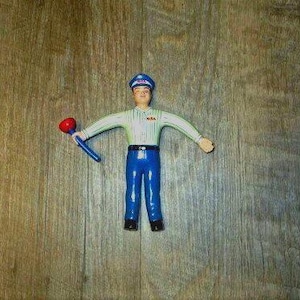 May include: A vintage toy figure of a man in a blue uniform, white shirt, and a blue cap. The figure is holding a red and blue object, possibly a plunger. The figure is standing on a wooden surface.