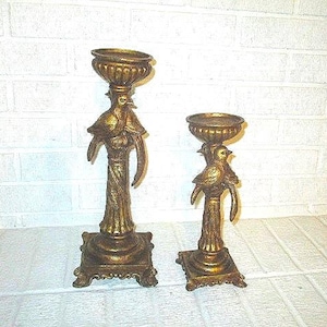 May include: Two antique gold-colored candle holders. The holders feature a bird design with a bowl-shaped top. The base is square with ornate detailing. The candle holders are different heights, with one taller than the other.