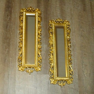 May include: Two ornate, rectangular gold mirrors with intricate scrollwork frames. The mirrors are vertically oriented and mounted on a light brown wooden surface. The frames have a detailed, vintage design.
