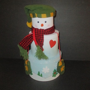May include: A stack of three round white and light blue gift boxes decorated to resemble a snowman. The top box features a snowman face, a green knit hat with a yellow pom-pom, and a red and black buffalo plaid scarf with green fringe ends.