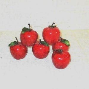 May include: A collection of six glossy red decorative apples arranged in a cluster. Each apple features a smooth, shiny surface, a dark brown stem at the top, and a small green leaf attached near the base of the stem, creating a uniform appearance.