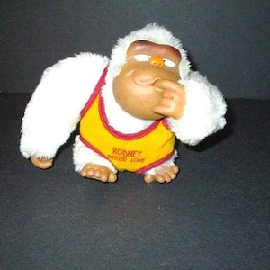 May include: A plush toy gorilla with white fur, a brown face, and a yellow vest with red trim. The vest has the text "RODNEY NEEDS LOVE". The gorilla is in a seated position, with its finger touching its lips.