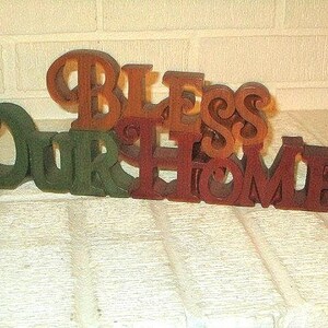 May include: Wooden sign with the words "BLESS OUR HOME" in varying shades of brown and green. The letters are cut out and have a rustic, three-dimensional appearance, suitable for home decor.
