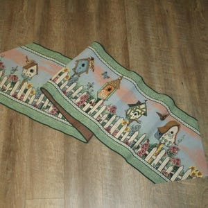 May include: Two decorative table runners with a whimsical birdhouse design. The runners feature a light blue background with birdhouses, flowers, and butterflies. A white picket fence borders the design, with a green border and tassels.