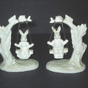 May include: Two white ceramic bunny figurines on swings. Each bunny is dressed in a detailed outfit and seated on a swing suspended from a tree-like structure. The base of each figurine is textured and detailed.