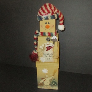 May include: A stacked wooden block snowman figurine wearing a red, white, and blue knit hat and scarf. The middle block features a sign reading Snowballs for Sale 3 cents. The bottom block displays a painted snowman with the text Let it Snow.