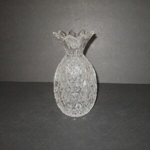 May include: A clear glass vase shaped like a pineapple, featuring a textured diamond pattern across its body and a scalloped, leaf-like rim at the top. The vessel is made of transparent, reflective glass with a distinct, repeating geometric design.