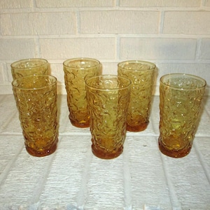 May include: A set of five amber-colored drinking glasses featuring a textured, organic surface pattern. Each glass has a cylindrical shape with a slightly wider rim and a solid base, showcasing a consistent golden-brown hue throughout the glass.