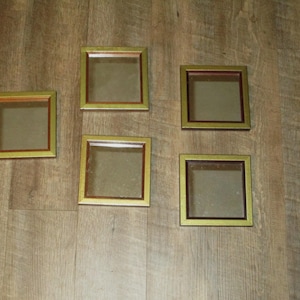 May include: Five square mirrors arranged in a group, each featuring a metallic gold outer frame and a thin dark red inner border surrounding the reflective glass center. The mirrors are identical in shape and design, displaying a clean, geometric look.