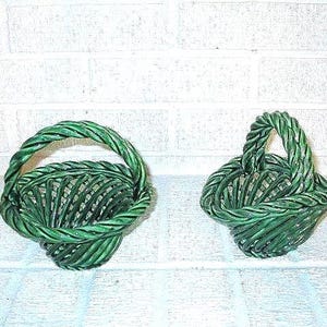 May include: Two green ceramic baskets with woven designs. The baskets have a twisted handle and a flared base. The baskets are a dark green color and appear to be handmade. The baskets are decorative and could be used for storage or display.