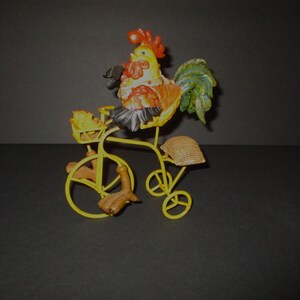 May include: A decorative figurine featuring a yellow, orange, and green rooster riding a yellow metal bicycle. The rooster has a red comb and wattle, while the bicycle includes a small front basket containing two small chicks and a tan seat.