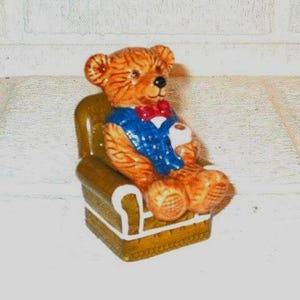 May include: A ceramic teddy bear figurine seated in a miniature armchair. The bear is brown with a blue vest and red bow tie, holding a white cup. The chair is brown with white trim.
