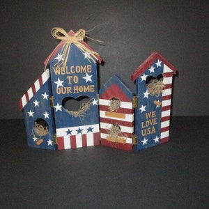May include: Three wooden birdhouses painted in red, white, and blue with patriotic themes. The birdhouses have star and stripe designs, with text that reads "WELCOME TO OUR HOME" and "WE LOVE USA". Each birdhouse has a heart-shaped opening and is decorated with straw.