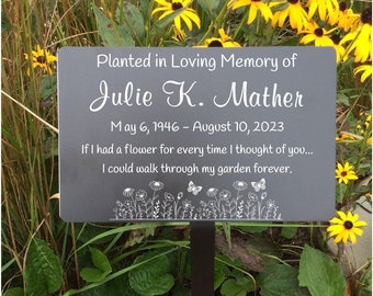 May include: A gray memorial garden sign with white text that reads "Planted in Loving Memory of Julie K. Mather May 6, 1946 - August 10, 2023 If I had a flower for every time I thought of you... I could walk through my garden forever." The sign is surrounded by yellow flowers and green foliage.