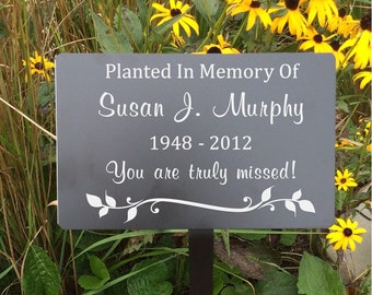 May include: A black metal garden sign with white text that reads "Planted In Memory Of Susan J. Murphy 1948 - 2012 You are truly missed!" The sign is surrounded by yellow flowers.