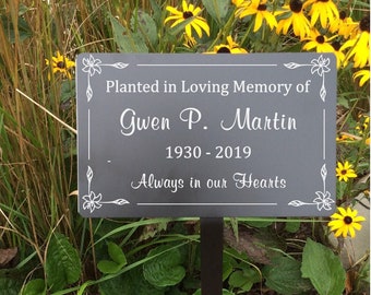 May include: A black metal garden marker with white text that reads "Planted in Loving Memory of Gwen P. Martin 1930-2019 Always in our Hearts". The marker is placed in a garden bed with yellow flowers.