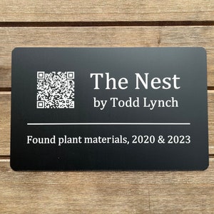 May include: Black rectangular sign with white text that reads "The Nest by Todd Lynch" and "Found plant materials, 2020 & 2023". A QR code is in the top left corner.