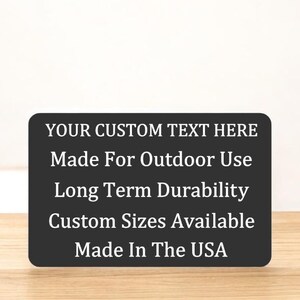 May include: Black rectangular sign with white text that reads "YOUR CUSTOM TEXT HERE", "Made For Outdoor Use", "Long Term Durability", "Custom Sizes Available", and "Made In The USA".