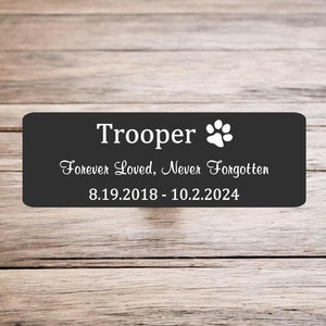 May include: Black rectangular plaque with white text that reads "Trooper" with a white paw print symbol. The text below reads "Forever Loved, Never Forgotten" and "8.19.2018 - 10.2.2024".