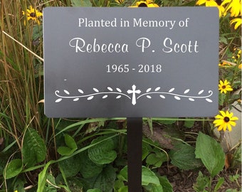 May include: A black metal garden marker with white text that reads "Planted in Memory of Rebecca P. Scott 1965 - 2018". The marker has a cross and a vine design. The marker is placed in a bed of yellow flowers.