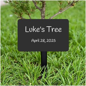 May include: Black sign with white text that reads "Luke's Tree" and "April 28, 2025". The sign is attached to a black metal stake and is planted in green grass.