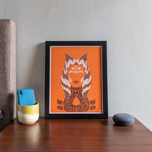 Op de afbeelding: Een ingelijst afdruk van een gestileerde illustratie van een personage uit Star Wars. Het personage is gemaakt van woorden die gerelateerd zijn aan het personage. De afdruk is op een oranje achtergrond en heeft een zwarte lijst.