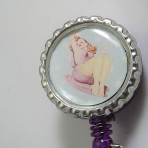 May include: A silver badge reel with a purple retractable cord and a round, silver cap with a vintage pin-up girl image.