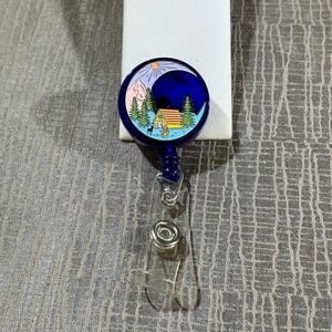 May include: A blue retractable badge reel with a nature scene. The design features a cabin, trees, mountains, and a sun and moon. The reel has a clear plastic strap and a metal clip.