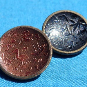 May include: Two antique buttons. One button is a reddish-brown color with an embossed design of a bird, boat, and other shapes. The other button is dark blue with a floral pattern. Both buttons have a gold-colored rim.