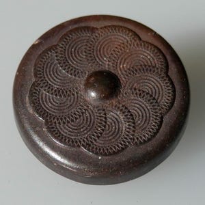 Könnte beinhalten: Ein dunkelbrauner Knopf mit einem erhabenen, kreisförmigen Design. Das Design zeigt einen zentralen Kreis, der von konzentrischen Kreisen umgeben ist, und einen gewellten Rand.