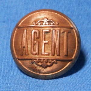 Könnte beinhalten: Ein runder, kupferfarbener Knopf mit dem Wort "AGENT" in erhabenen Buchstaben. Der Knopf hat ein dekoratives Design am Rand.