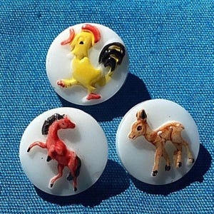 Puede incluir: Tres botones redondos blancos con ilustraciones de animales coloridos. El primer botón presenta un gallo amarillo con cola negra y cresta roja. El segundo botón presenta un caballo rojo con crin y cola negras. El tercer botón presenta un ciervo marrón con manchas blancas.