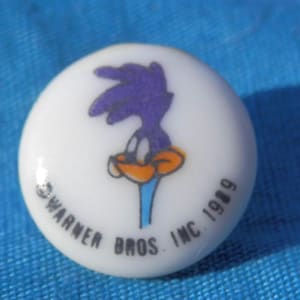 Puede incluir: Un botón de cerámica blanca con una imagen de dibujos animados de Correcaminos, un pájaro morado y naranja, con el texto "Warner Bros. Inc. 1989".