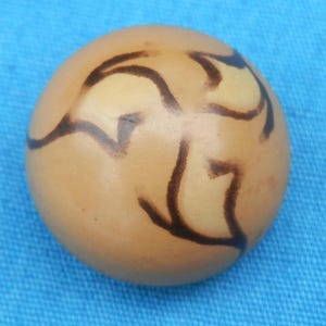 May include: A round, tan-colored ceramic ornament with a black abstract design. The design resembles a stylized bird or flower.