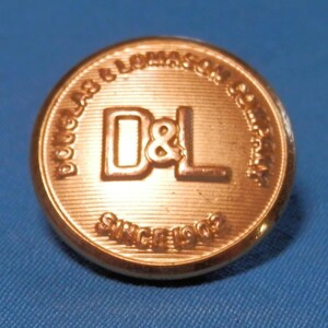 Könnte beinhalten: Ein runder, kupferfarbener Knopf mit dem geprägten Text "DOUGLAS & LOMASON COMPANY" am Rand. In der Mitte befinden sich die Buchstaben "D&L" und der Text "SINCE 1902". Der Knopf ist vor einem blauen Hintergrund platziert.