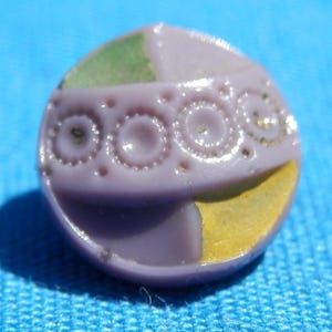 May include: A purple glass button with a yellow, green, and white design. The button has a raised, textured pattern.