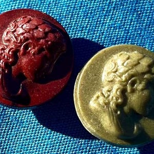 May include: Two round, decorative buttons with a raised image of a woman's profile. The button on the left is a deep red color, while the button on the right is a light brown color.