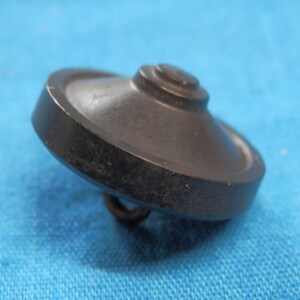 Könnte beinhalten: Nahaufnahme eines dunkelbraunen Knopfes mit kreisförmiger, leicht gewölbter Form. Der Knopf hat eine kleine, erhabene Mitte und eine Metallschlaufe auf der Unterseite zum Annähen. Der Knopf ist vor blauem Hintergrund platziert.