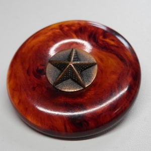 May include: A round, brown and orange-swirled object with a bronze star in the center. The star is raised and has a textured surface. The object appears to be a decorative knob or button.