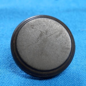 Könnte beinhalten: Ein runder, antiker Metallknopf mit einem dunklen, strukturierten Rand und einer helleren, abgenutzten Mitte. Der Knopf ist wahrscheinlich aus Stahl oder einem ähnlichen Metall gefertigt und weist Gebrauchsspuren auf. Der Knopf ist vor blauem Hintergrund platziert.